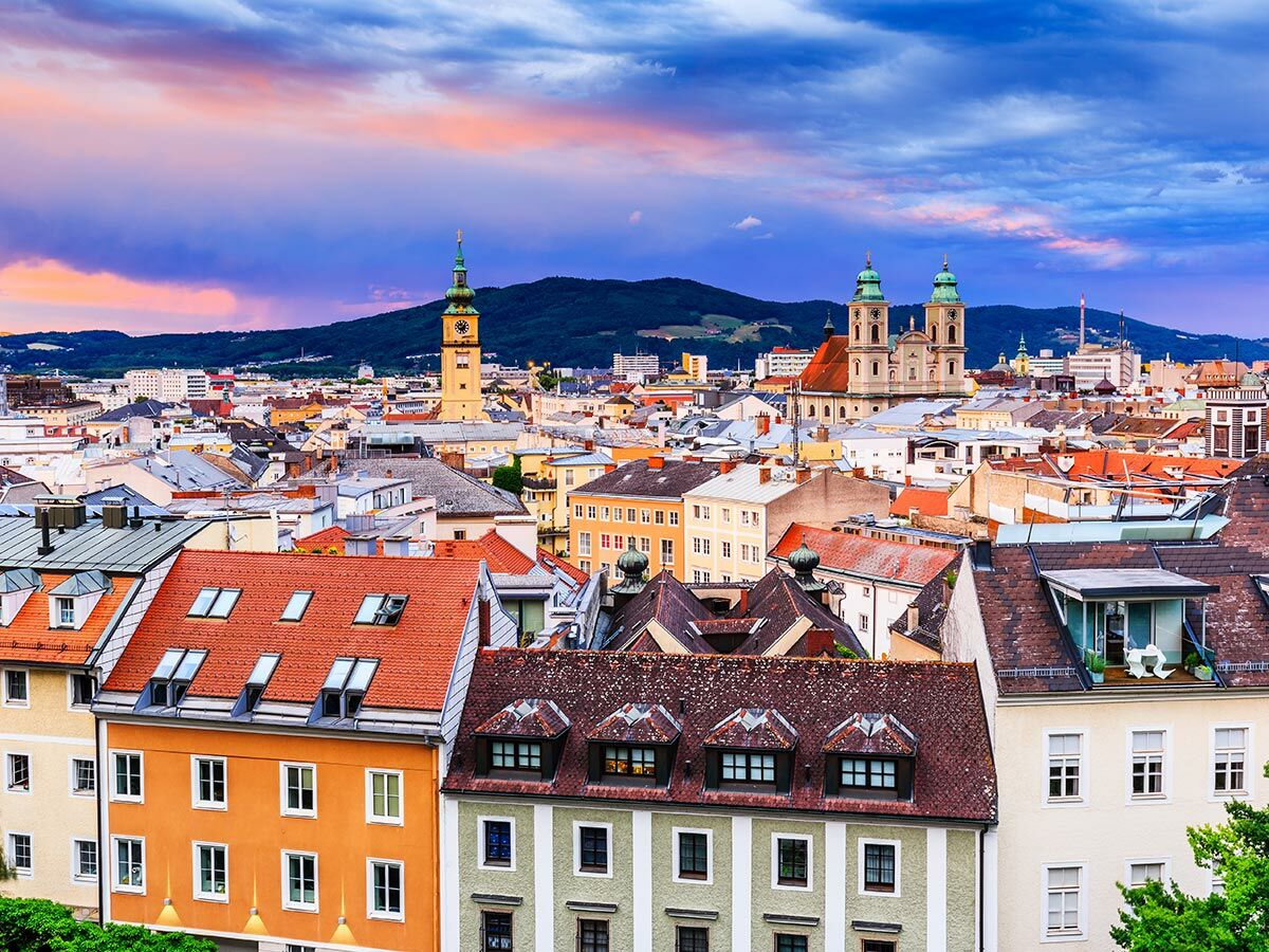 Malerisches Panorama der Altstadt von Linz, Österreich, bei Sonnenuntergang mit farbenfrohem Himmel, historischen Gebäuden, Kirchen und Bergen.