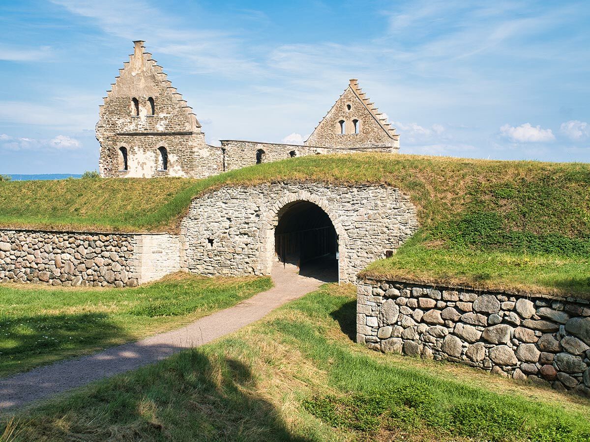 Eketorp Festung auf Öland, Schweden: mittelalterliche Burgruine mit Steintor, grasbewachsenen Wällen und historischen Gebäuden unter blauem Himmel.