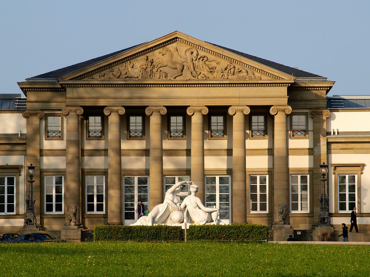 Fassade des Neuen Schlosses in Stuttgart mit ionischen Säulen, Reliefgiebel und weißen Marmorskulpturen im Vordergrund.