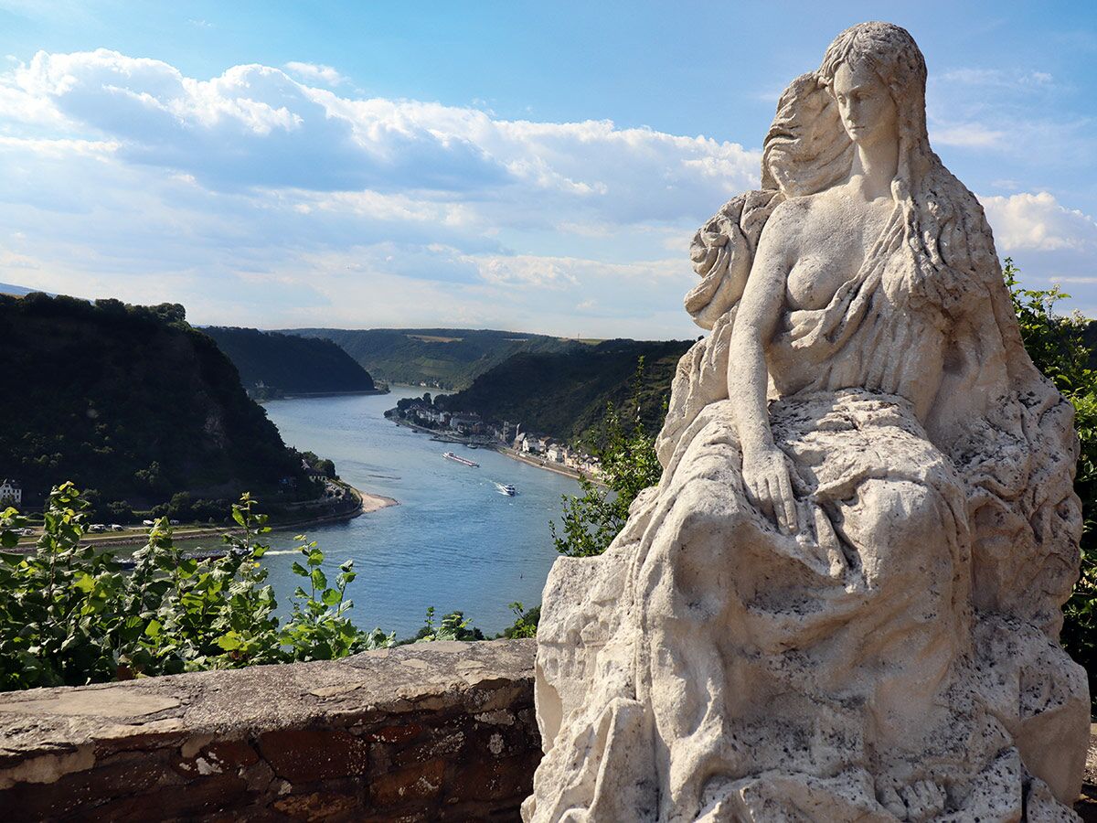 Loreley-Statue blickt über das malerische Rheintal mit Fluss, grünen Hügeln, Dorf und Schiffsverkehr am sonnigen Tag.