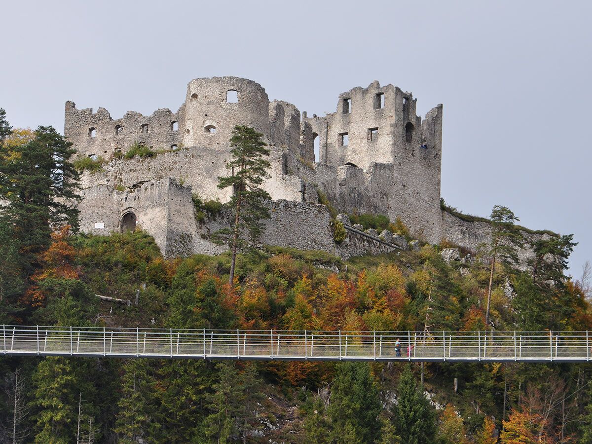 Im Herbst ragt die Burgruine Ehrenberg über die Highline179 Hängebrücke, auf der zwei Personen im bunten Wald spazieren.