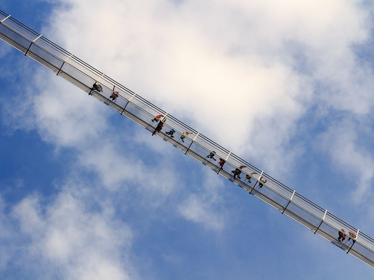 Von unten aufgenommener Blick auf eine moderne Glas-Hängebrücke mit mehreren Menschen, die unter blauem Himmel und weißen Wolken entlanglaufen.