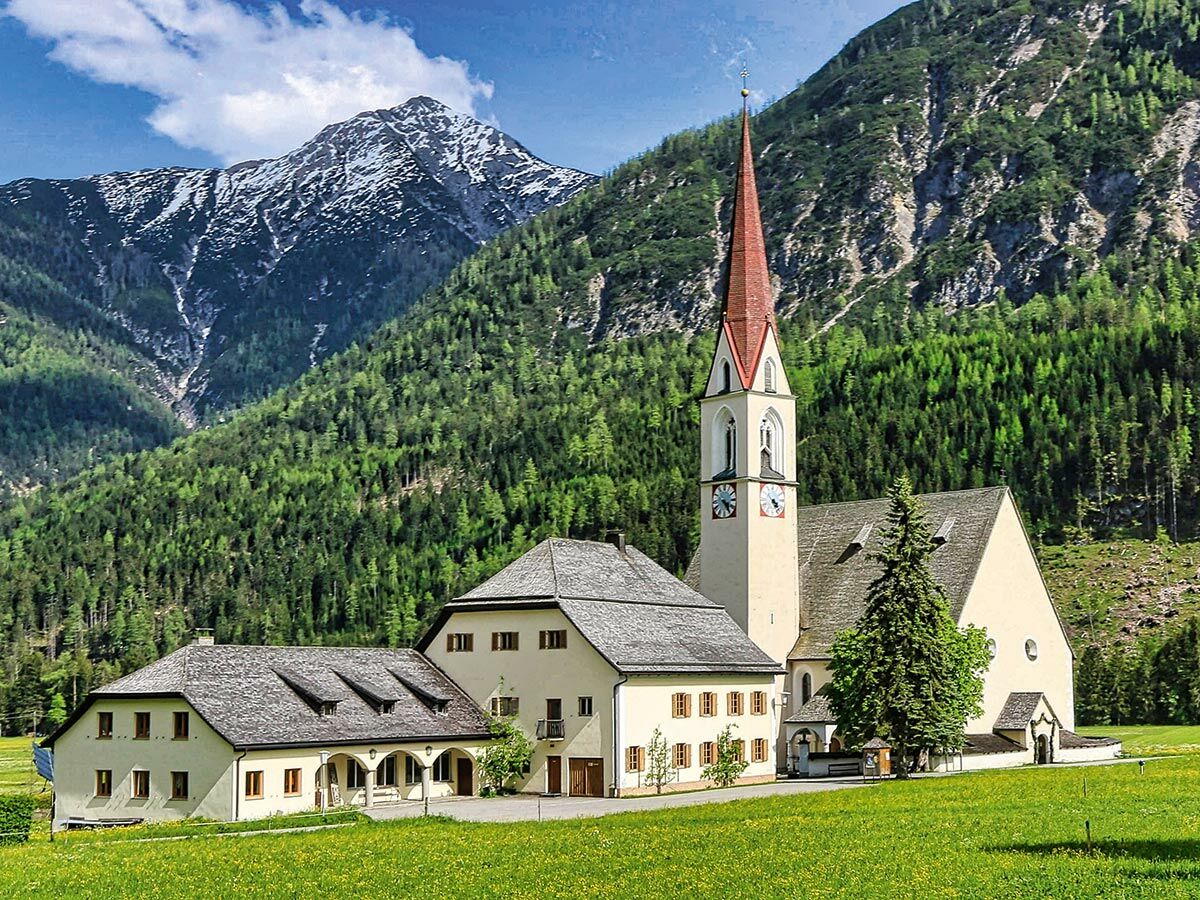 Blick auf eine historische Alpenkirche mit rotem Turm und angrenzenden Gebäuden vor grünen Wäldern und einem schneebedeckten Berg.