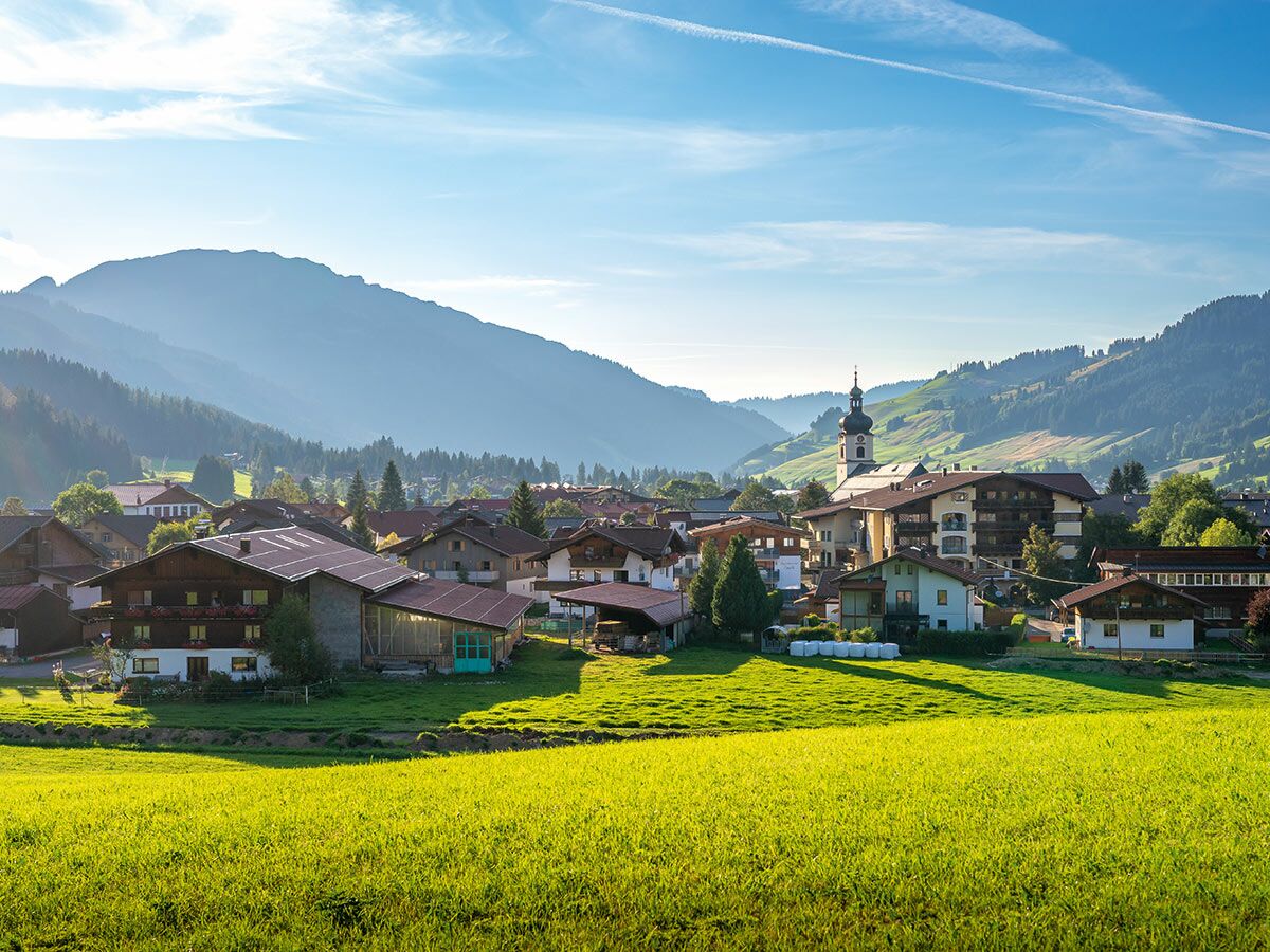 Blick auf ein idyllisches Bergdorf mit traditionellen Häusern, Kirchturm und grünen Wiesen vor majestätischen Alpen.