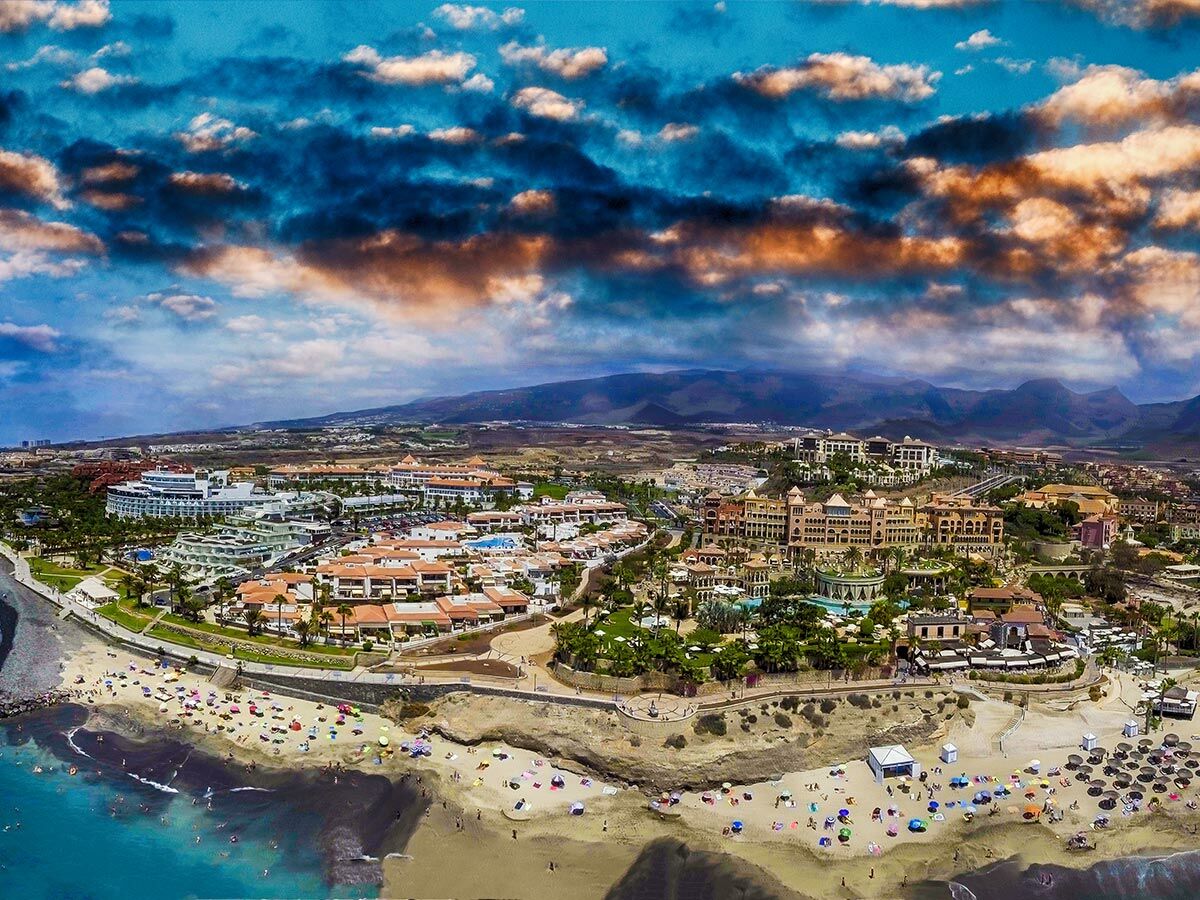 Luftaufnahme von luxuriösen Hotels und belebten Sandstränden mit vielen Sonnenschirmen an der Küste Teneriffas unter dramatischem Himmel.