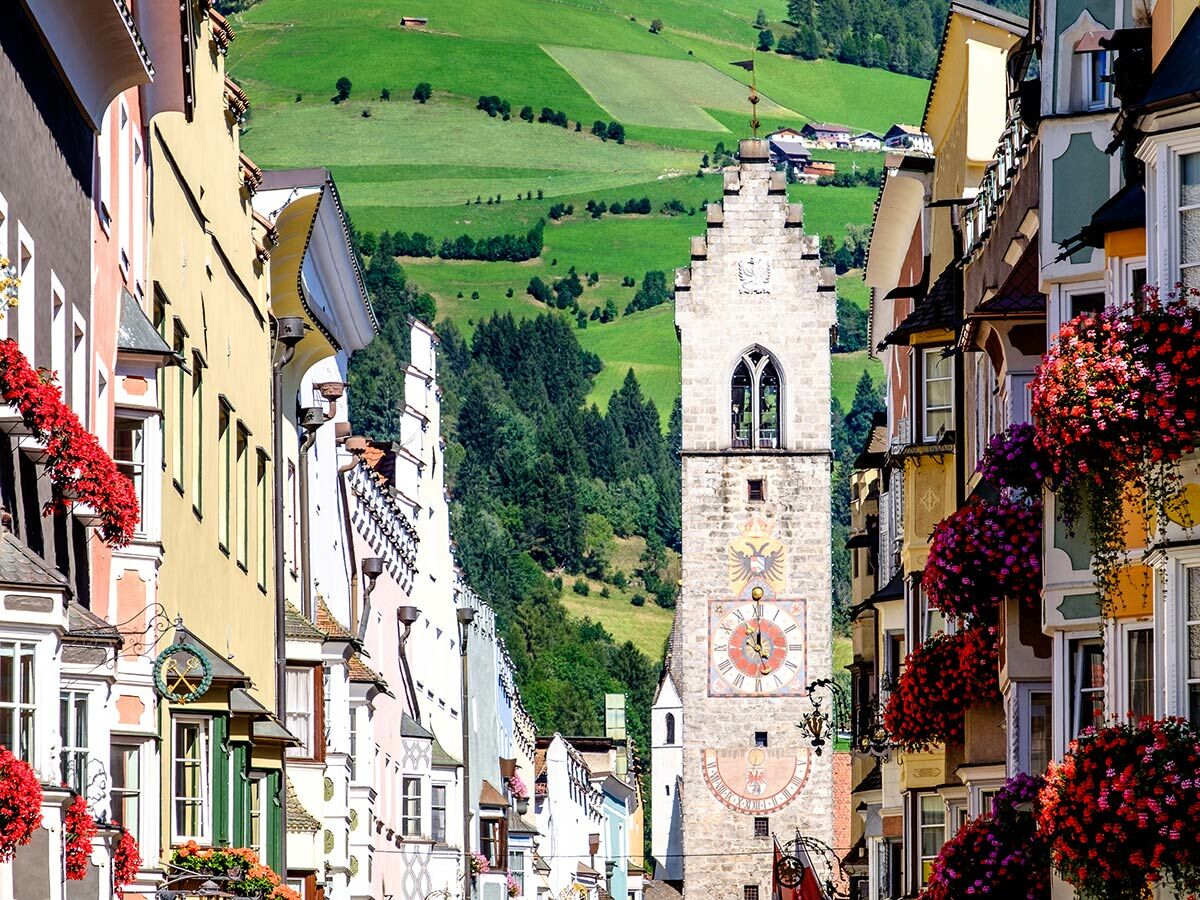 Historische Alpenstadt mit farbenfrohen Häusern, blühenden Blumenkästen und einem markanten Glockenturm, umgeben von grünen Hügeln.