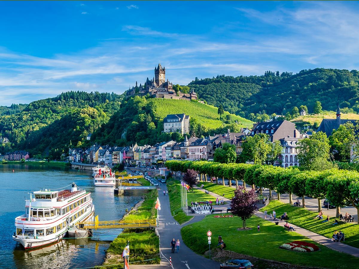 Panoramablick auf die Reichsburg Cochem an der Mosel mit malerischer Altstadt, Flusskreuzfahrtschiffen und grünen Weinbergen.