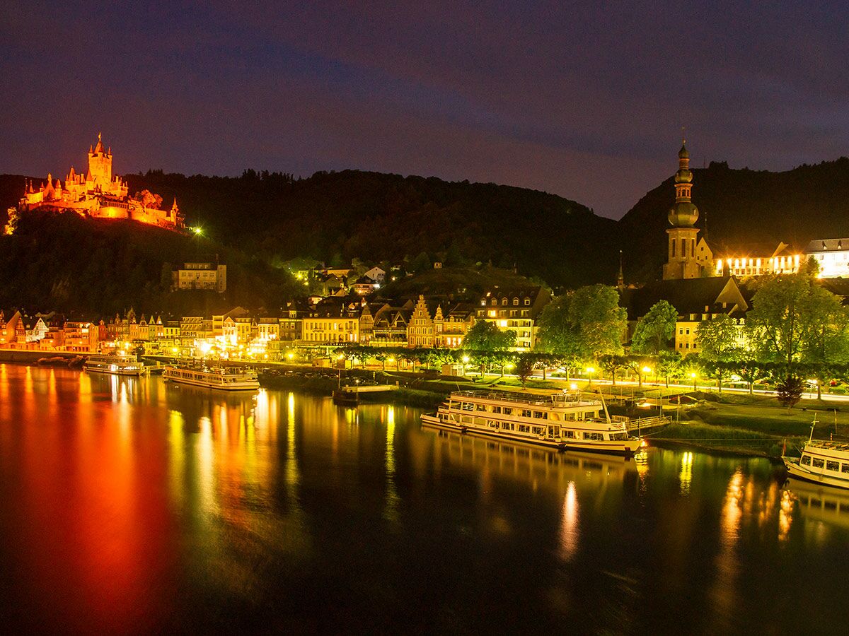 Panoramablick auf Cochem bei Nacht mit beleuchteter Reichsburg, Mosel, Schiffen und Stadtgebäuden.