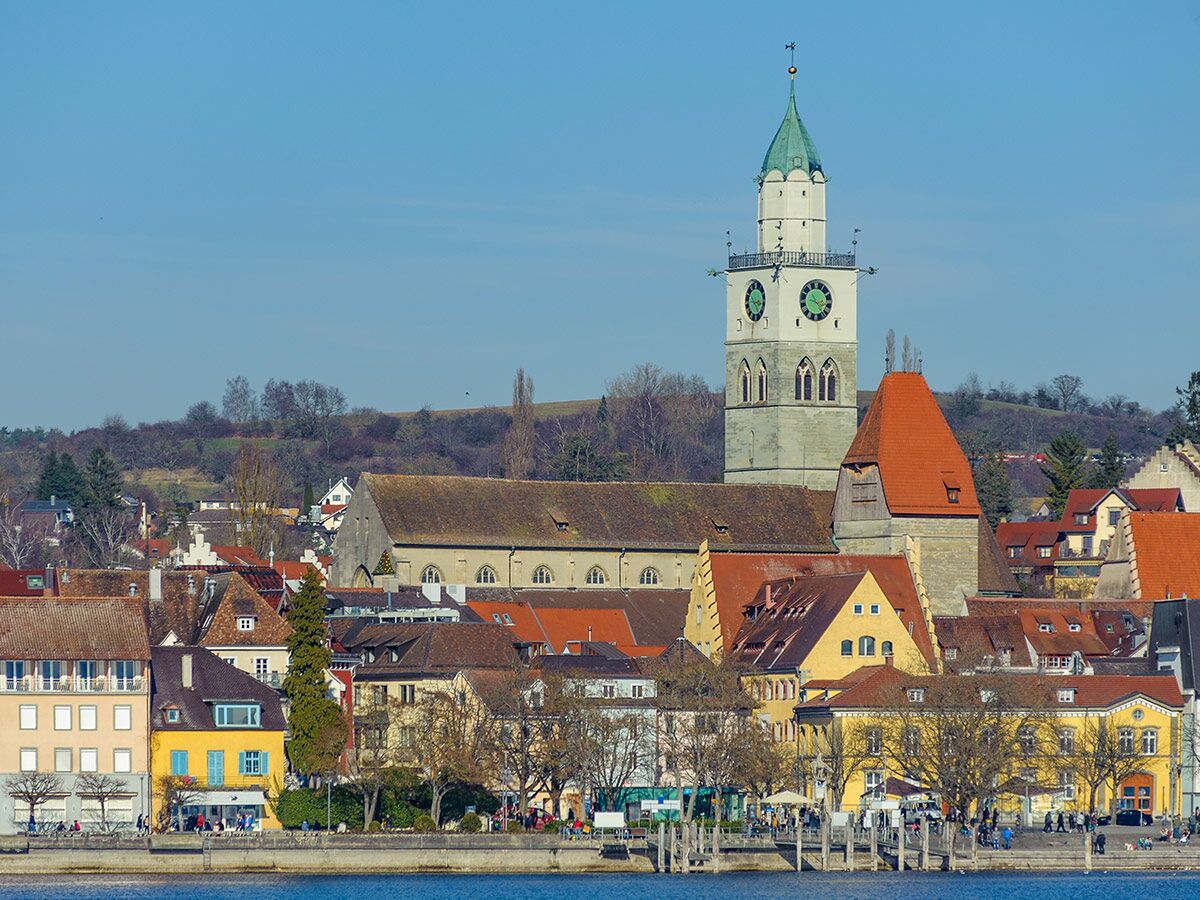 Blick auf Radolfzell am Bodensee mit dem Münster, der Altstadt und dem belebten Uferbereich unter blauem Himmel.