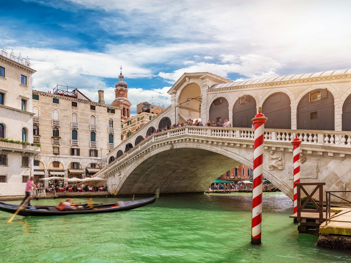 Panoramablick auf die Rialtobrücke in Venedig mit einer Gondel auf dem Canal Grande, historischen Gebäuden und Touristen.