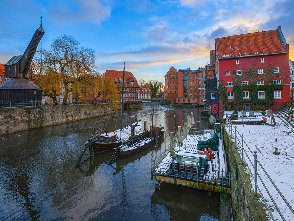 Winterliche Stadtansicht Lüneburgs mit dem Alten Kran am Kanal, historischen Gebäuden, schneebedeckten Booten und Stegen bei Sonnenuntergang.