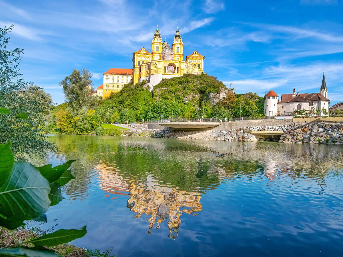Das imposante Stift Melk in Österreich thront auf einem Hügel und spiegelt sich malerisch im ruhigen Fluss unter blauem Himmel.