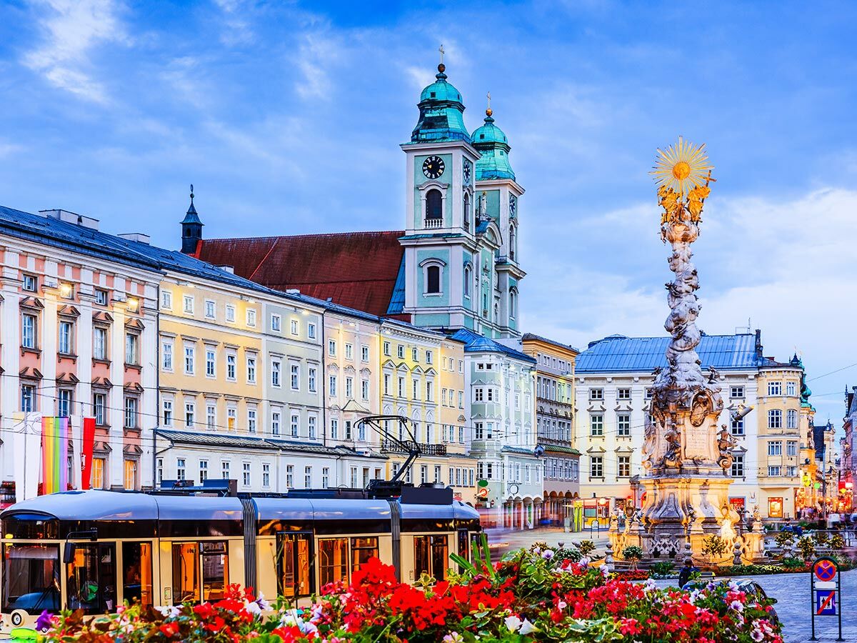 Abendaufnahme des Linzer Hauptplatzes mit beleuchteter Dreifaltigkeitssäule, Alter Dom, bunten Häusern und einer Straßenbahn.