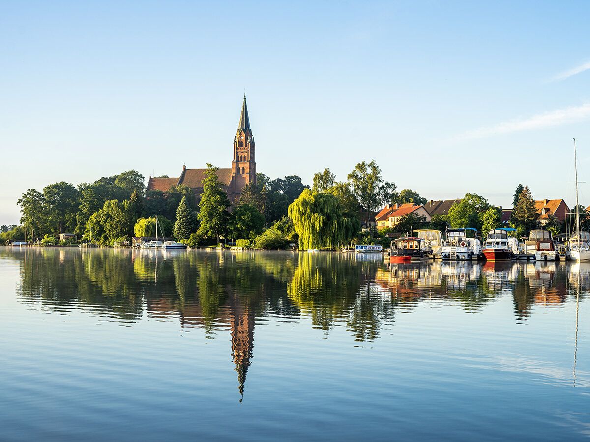 Idyllische Seeufer-Szene mit roter Dorfkirche, grünen Bäumen und reflektierenden Booten in ruhigem Wasser bei Sonnenschein.