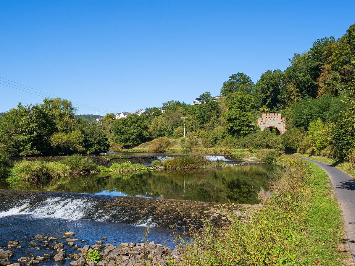 Idyllische Flusslandschaft mit sanften Wehren, umgeben von grünen Wäldern. Ein Radweg führt entlang des Ufers, im Hintergrund eine historische Bogenbrücke.