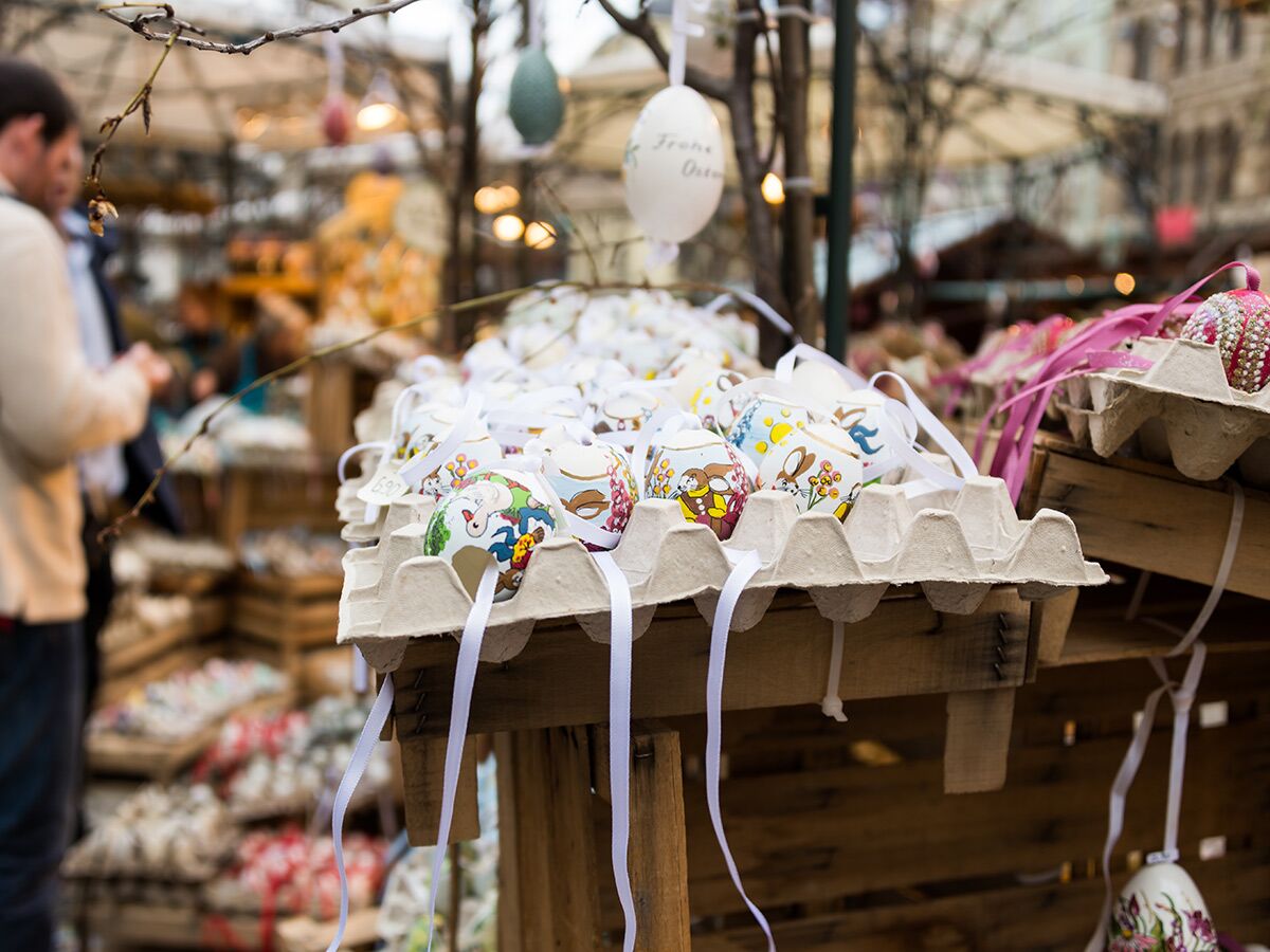 Nahaufnahme von bunten, handbemalten Ostereiern in Eierkartons auf einem Ostermarkt, mit Käufern im Hintergrund.