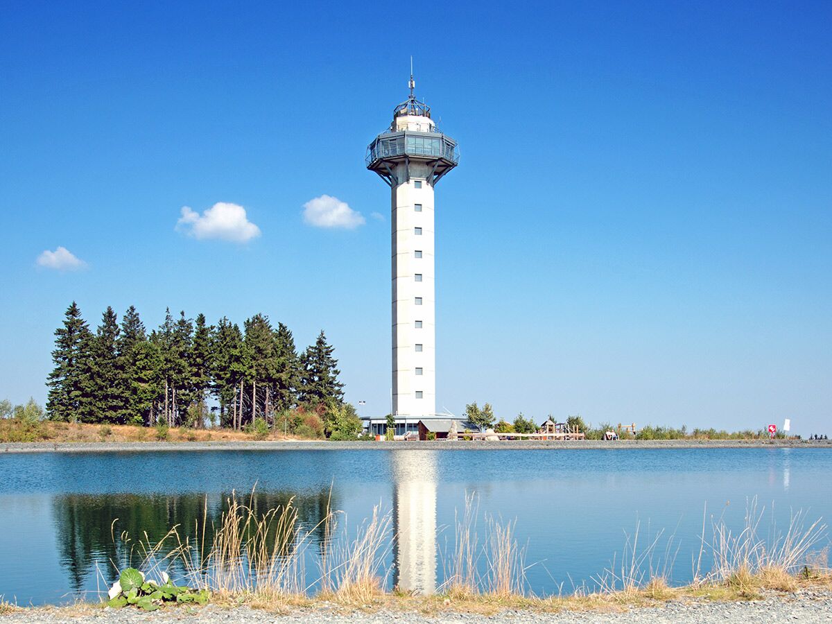 Hoher Aussichtsturm mit spiegelndem Wasser und Tannenbäumen unter blauem Himmel, ideal für Wander- und Freizeitaktivitäten.