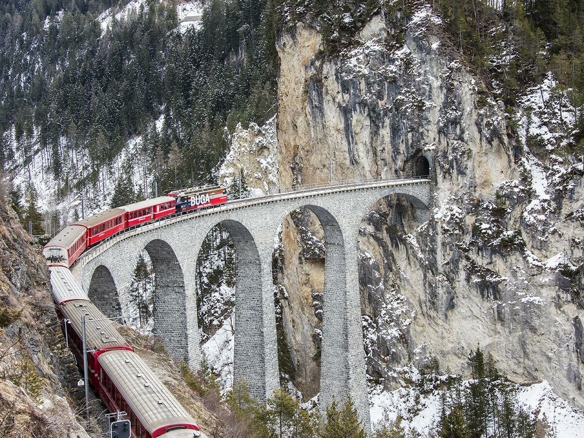 Roter RhB Zug fährt über den Landwasser Viadukt in verschneiter Berglandschaft zu einem Tunnel.