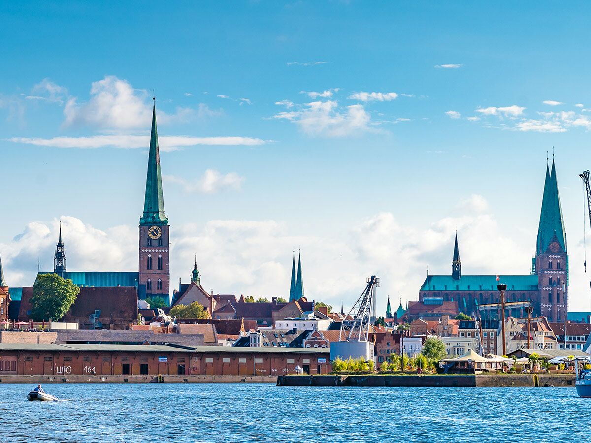 Historisches Stadtpanorama von Lübeck mit St. Petri, Marienkirche und Dom; Flussufer mit Lagerhäusern und Boot.
