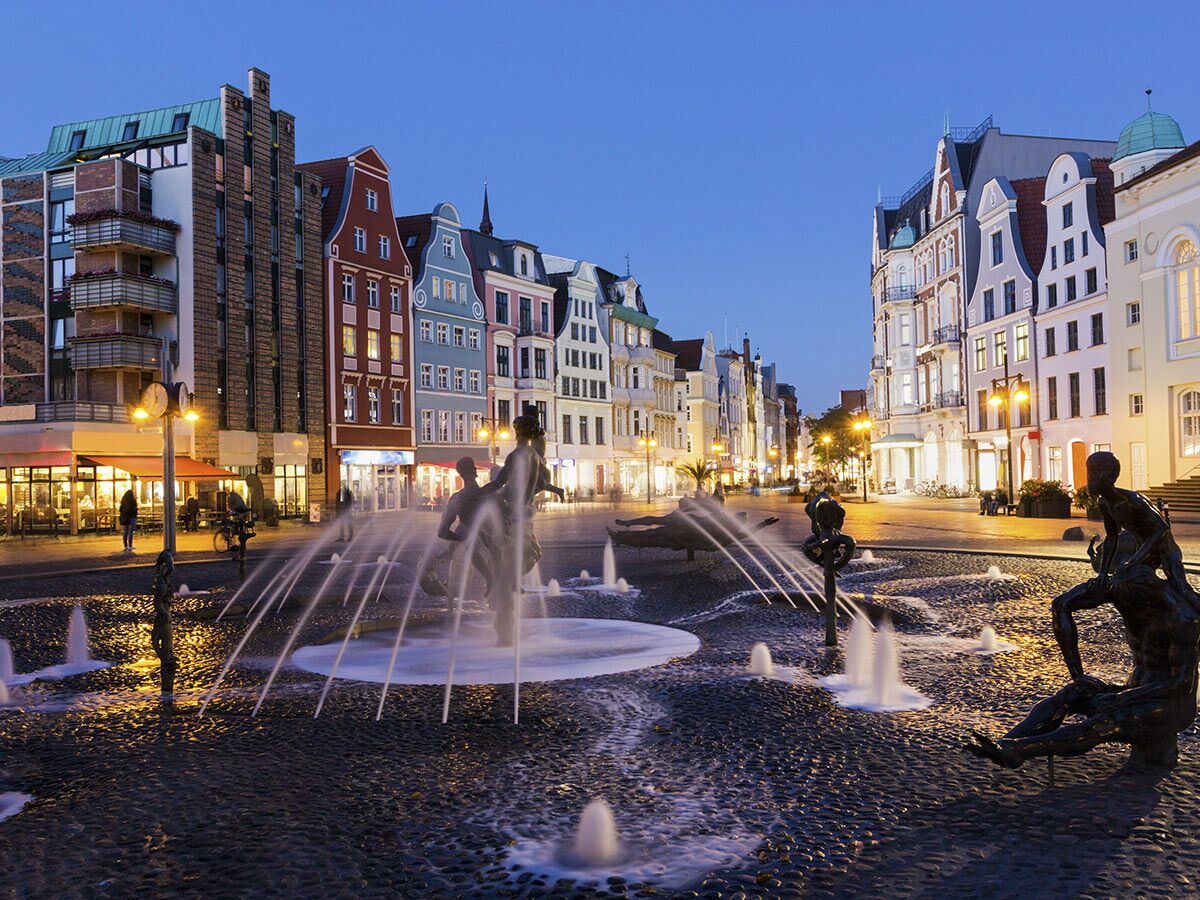 Brunnen der Lebensfreude auf dem Neuen Markt in Rostock bei Abenddämmerung, umgeben von beleuchteten Giebelhäusern.