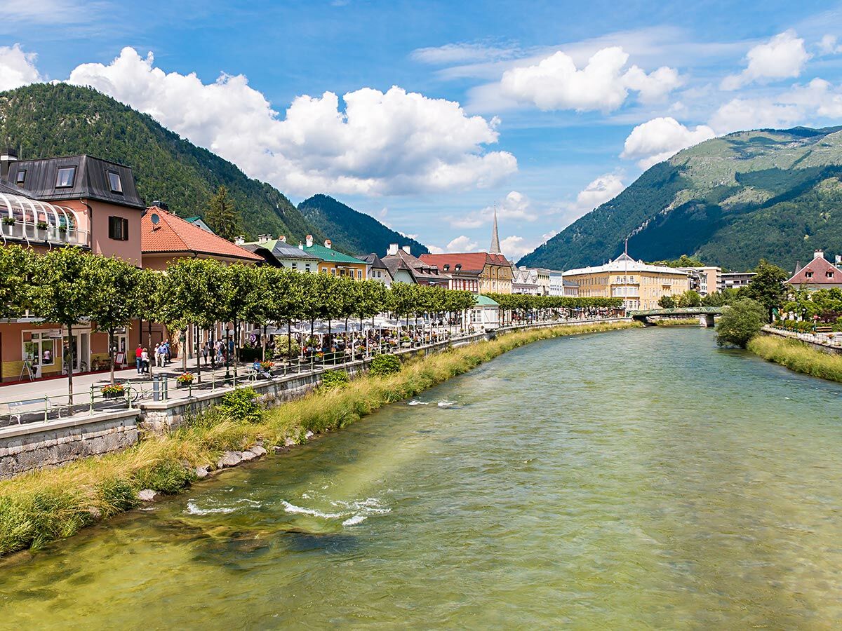 Blick auf Bad Ischl mit der Traun, gesäumt von historischen Gebäuden, Cafés und belebter Promenade vor alpiner Bergkulisse.
