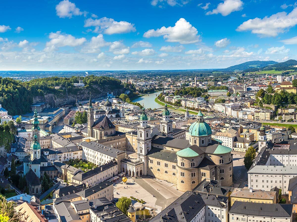 Panoramablick auf Salzburg mit Salzach, Altstadt, Dom und Festung Hohensalzburg unter blauem Himmel mit Wolken.