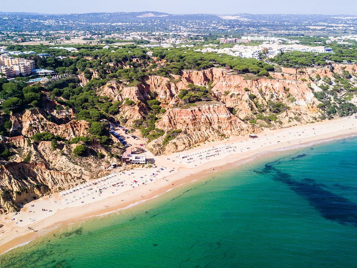 Luftaufnahme von Praia da Falésia, Algarve. Rote Klippen, grüne Bäume, goldener Sandstrand, türkisblaues Meer. Viele Badegäste.