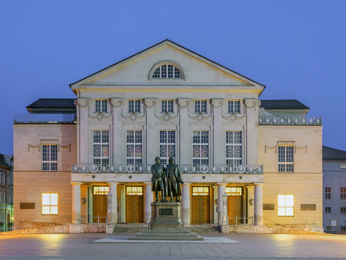 Deutsches Nationaltheater Weimar bei Dämmerung mit Goethe- und Schiller-Denkmal und beleuchteten Fenstern.