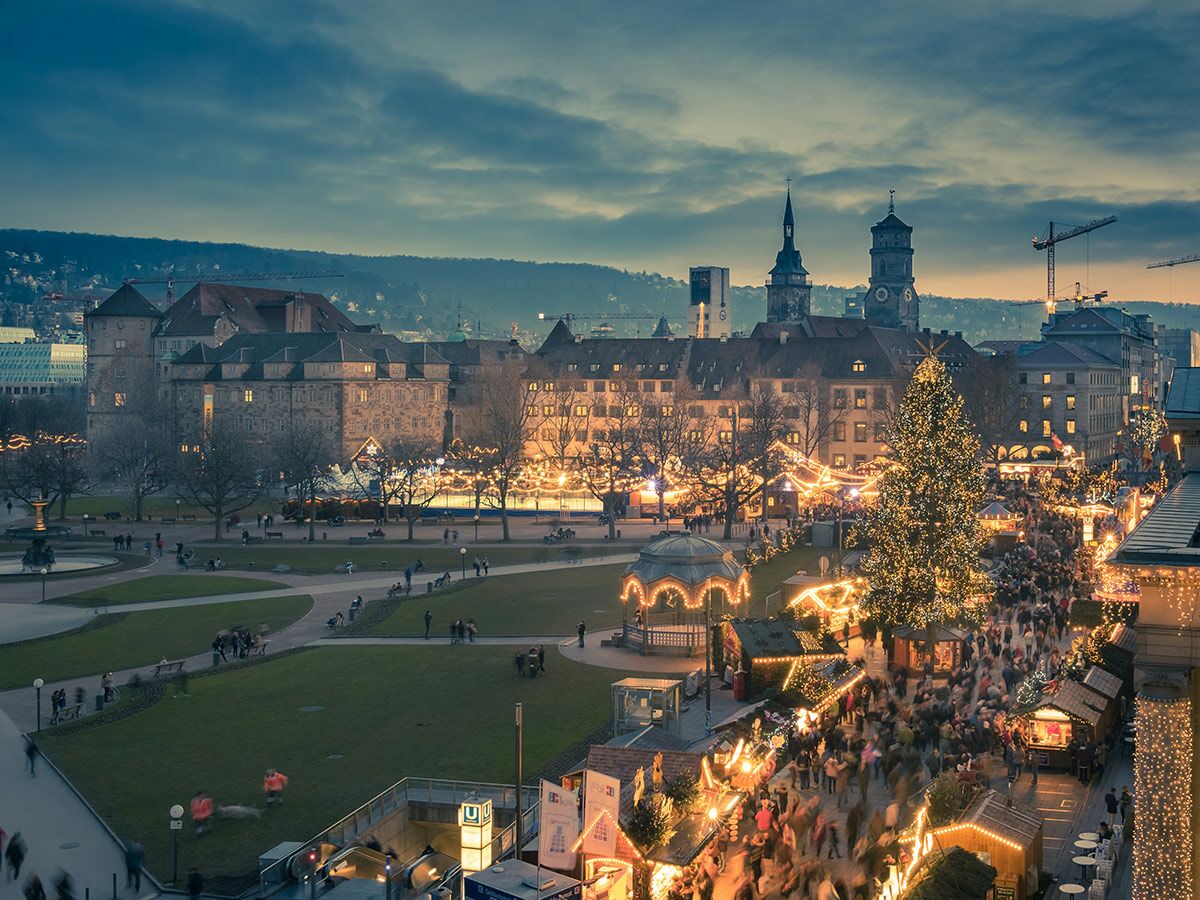 Beleuchteter Weihnachtsmarkt am Schlossplatz in Stuttgart bei Abenddämmerung mit Tannenbaum, Buden und vielen Menschen.