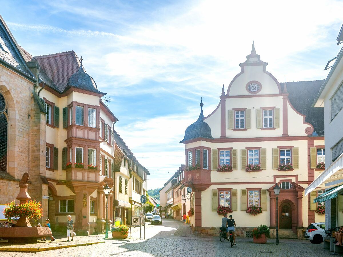 Sonnenbeschienene historische Gasse in einer deutschen Altstadt mit Kopfsteinpflaster, farbigen Häusern, Menschen und Brunnen.