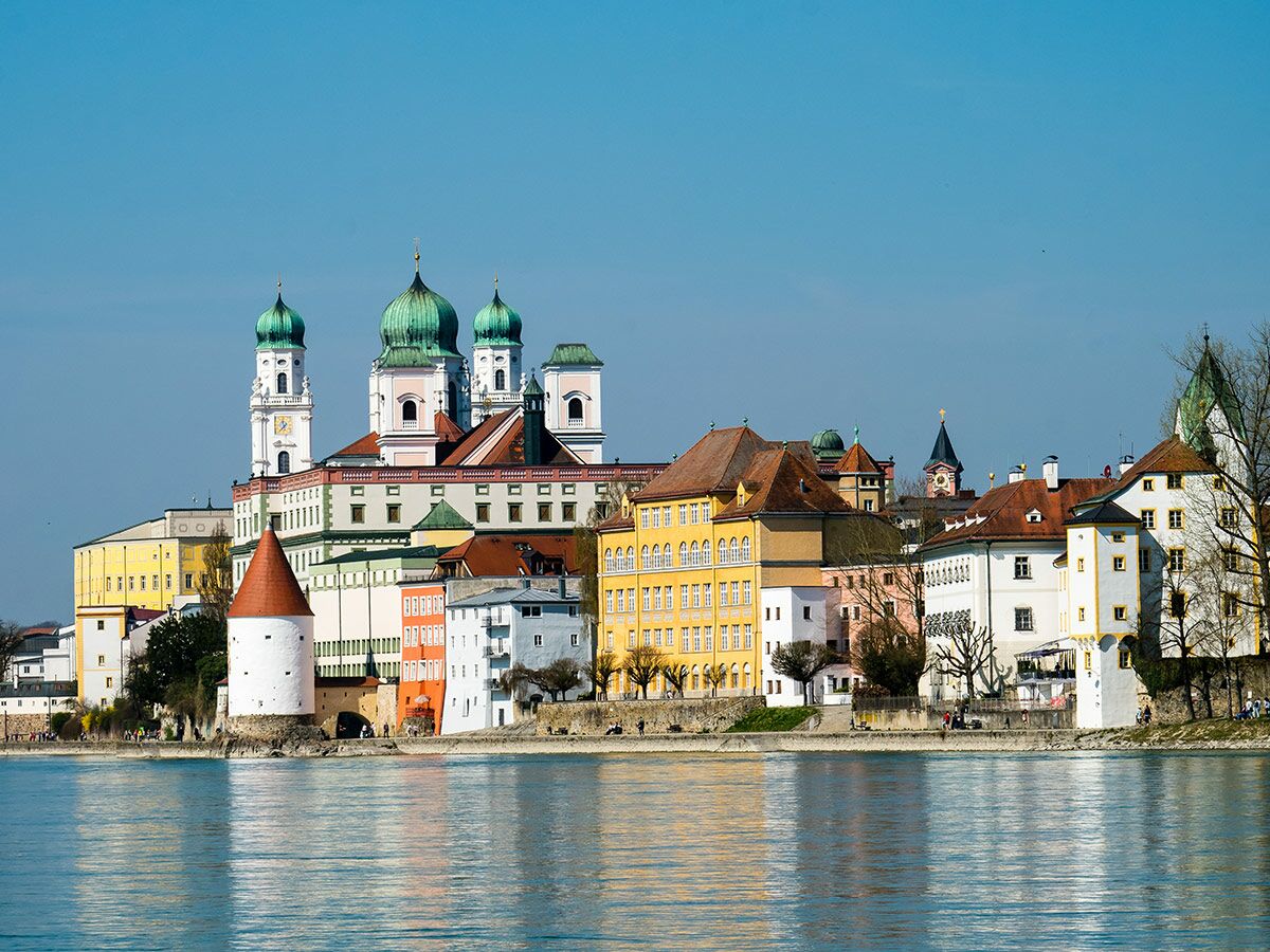 Panorama von Passau mit Dom St. Stephan und farbigen Häusern am Flussufer, strahlend blauem Himmel und reflektierendem Wasser.