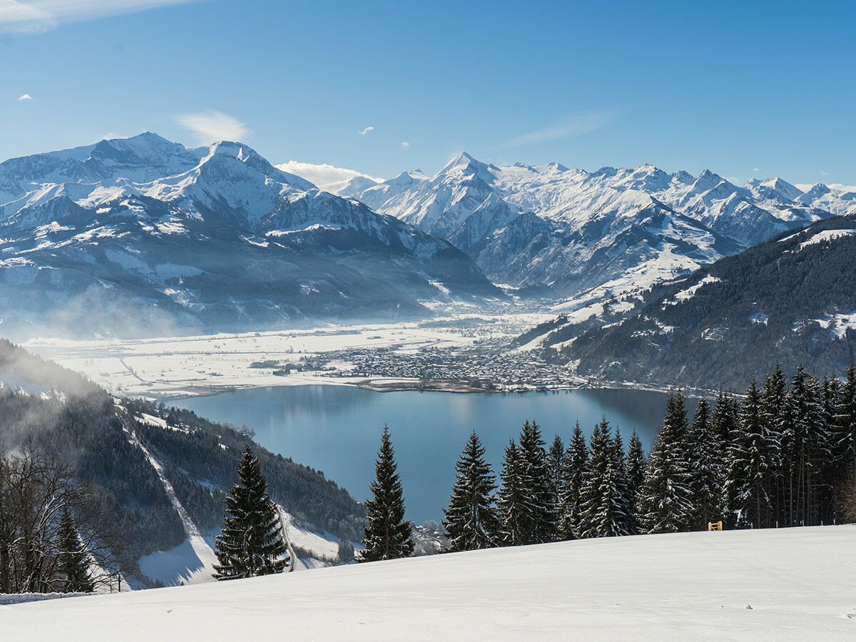 Panorama einer idyllischen Winterlandschaft mit schneebedeckten Alpen, einem tiefblauen See und einem Dorf an dessen Ufer unter klarem Himmel.