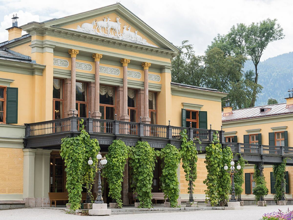 Gelbes historisches Schloss mit klassischer Architektur, Säulen, Hirsch-Relief, rankenbewachsenem Balkon und Bergkulisse.