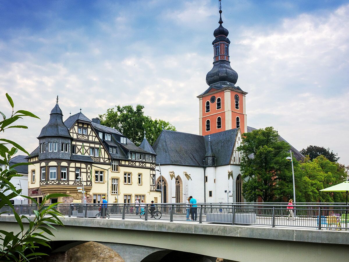 Blick auf eine deutsche Stadt mit historischer Kirche (Zwiebelturm), Fachwerkhaus und Menschen auf einer modernen Brücke an einem bewölkten Tag.