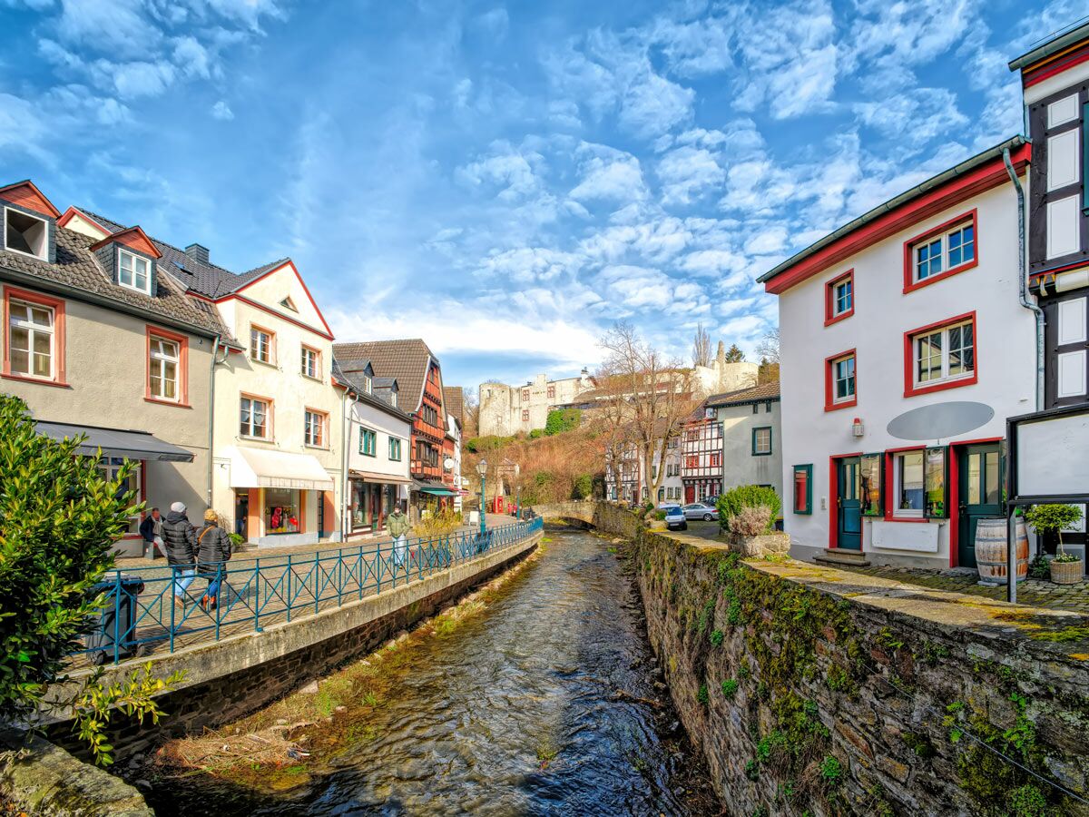 Malerische historische Altstadt in Europa mit Fluss, bunten Gebäuden, Fachwerkhäusern, Menschen und Burgruine am Hügel unter blauem Himmel.