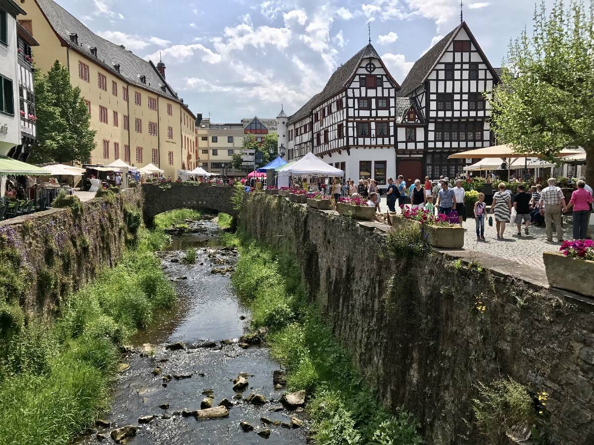 Malerische deutsche Altstadt mit Fachwerkhaus-Architektur, einem Bach, einer Steinbrücke und einem belebten Markt unter blauem Himmel.