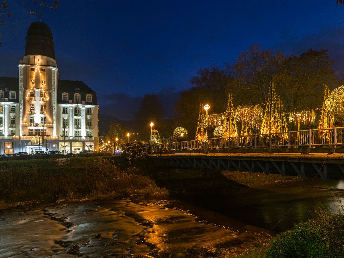 Nachtaufnahme einer Brücke mit goldener Weihnachtsbeleuchtung und einem hell erleuchteten Hotel am Fluss.