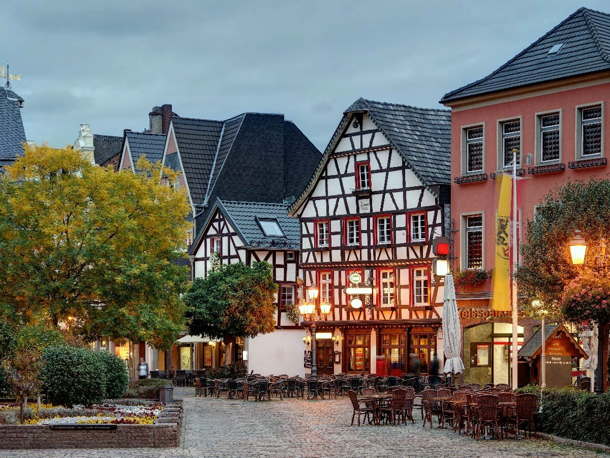 Historischer Marktplatz mit Fachwerkhäusern, Restaurants, Tischen, Stühlen und beleuchteten Laternen bei Abenddämmerung.