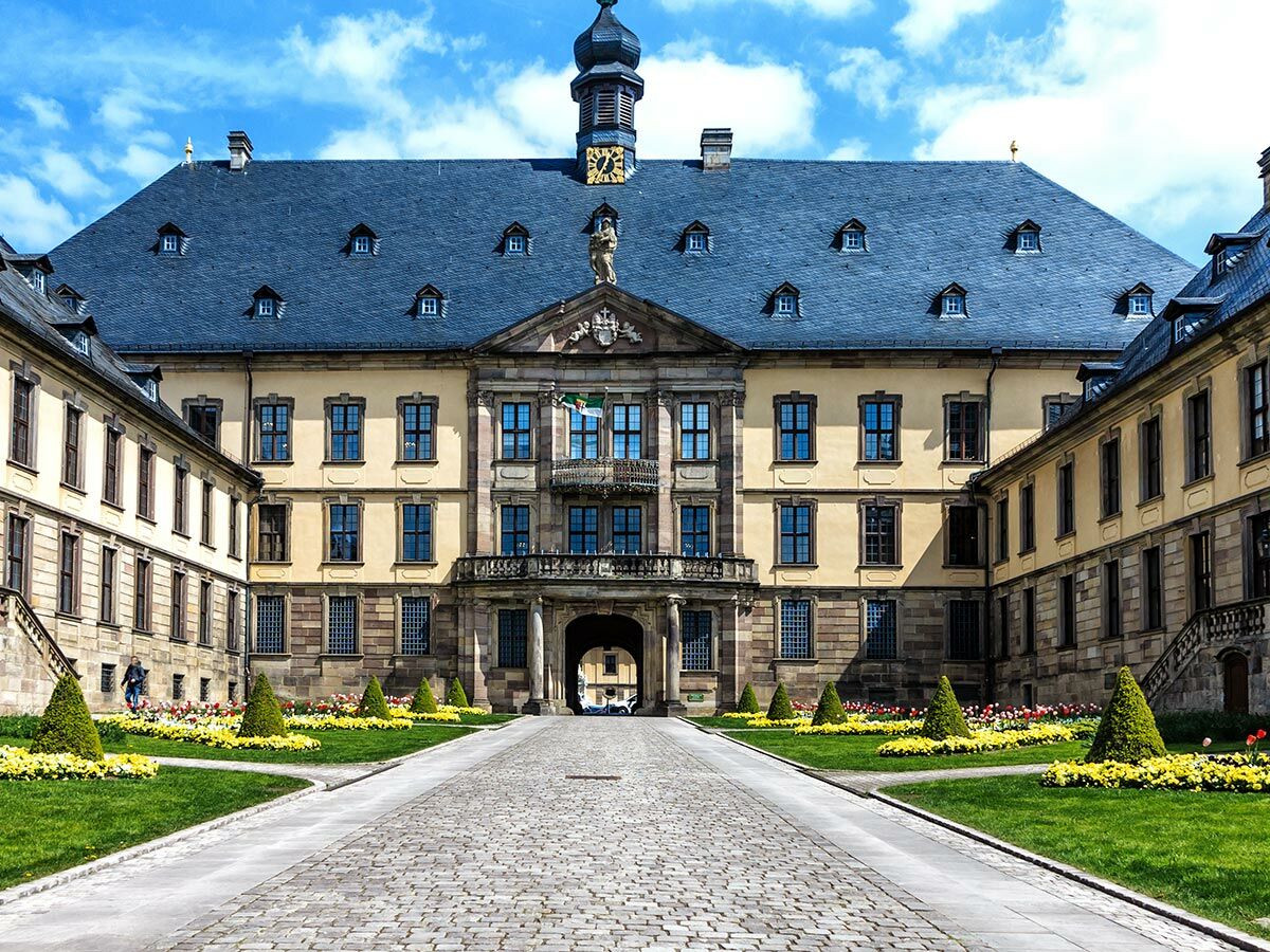 Großes Barockschloss mit gelber Fassade, blauem Dach, Uhrturm, Garten und gepflastertem Weg unter blauem Himmel.