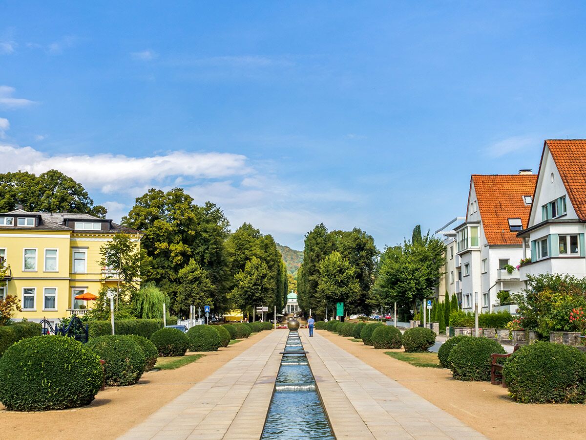 Weitläufige, grüne Promenade mit zentralem Wasserlauf, gesäumt von Bäumen und Gebäuden unter blauem Himmel.