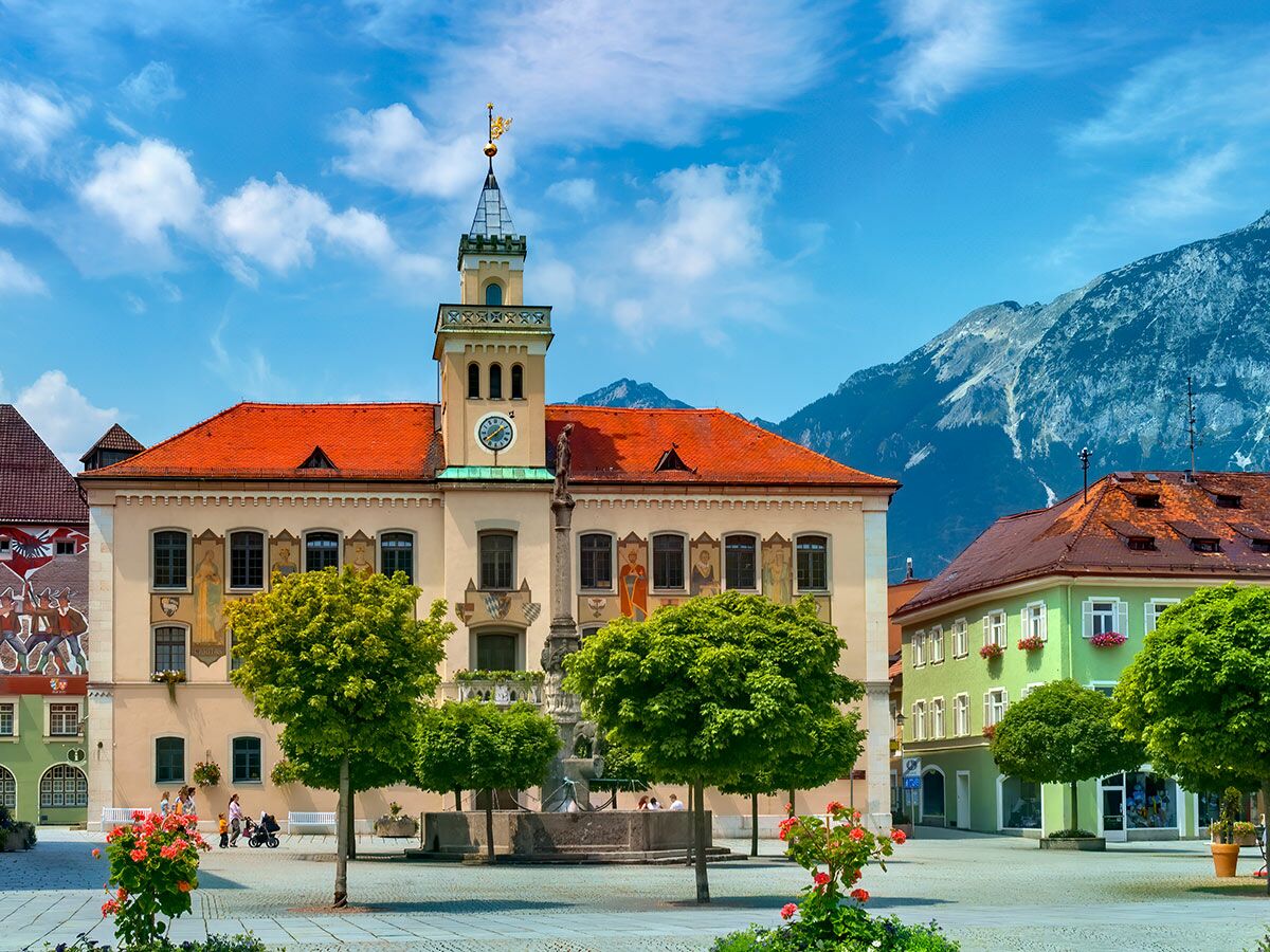 Belebter Marktplatz mit historischem Rathaus, Uhrturm, Brunnen und Bergen unter blauem Himmel in einer malerischen Stadt.