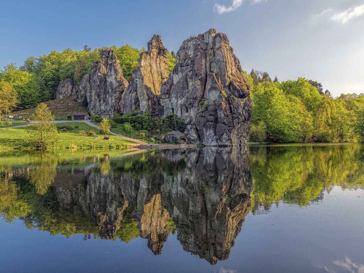 Die Externsteine, eine beeindruckende Felsformation, spiegeln sich im ruhigen See, umgeben von grünem Wald und blauem Himmel an einem sonnigen Tag.