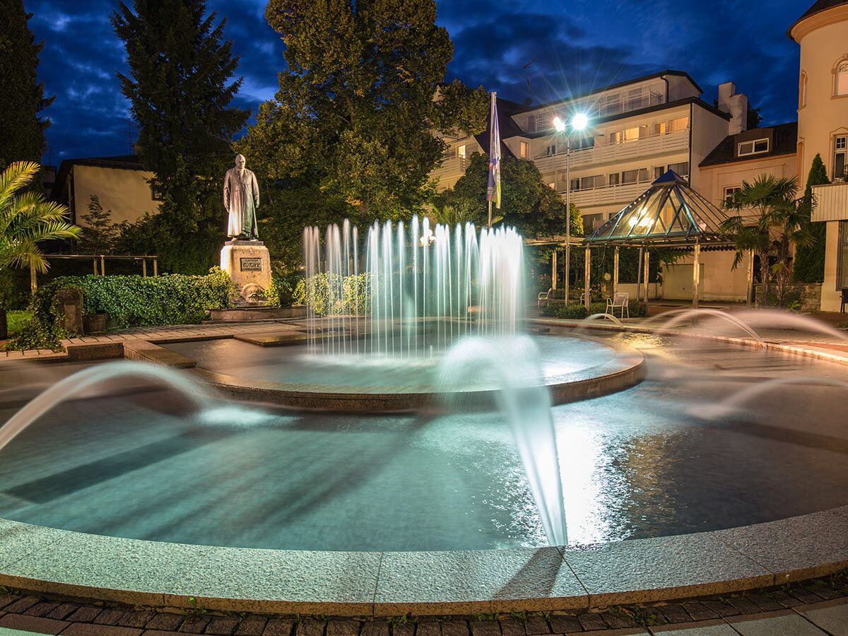 Nächtliche Ansicht eines beleuchteten Parks mit großem Brunnen, Wasserspielen, Statue eines Mannes, Bäumen und Gebäuden im Hintergrund.