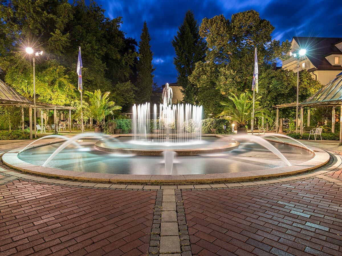Nachts beleuchteter Brunnen in einem Park mit Wasserspielen, einer Statue, Bäumen und Sitzgelegenheiten auf einem gepflasterten Platz.
