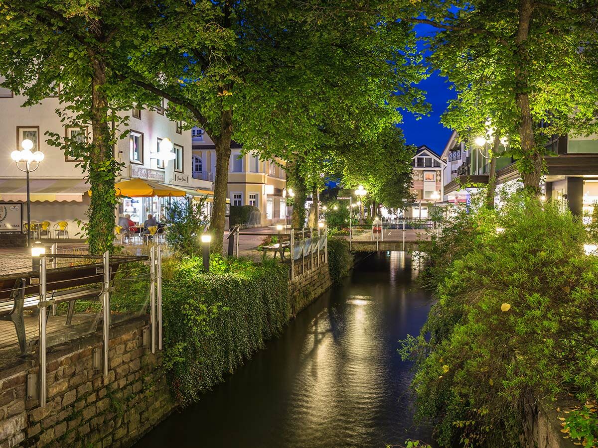 Beleuchteter Stadtkanal bei Nacht mit grünen Bäumen, alten Gebäuden und gemütlichen Cafés am Ufer, romantische Atmosphäre.