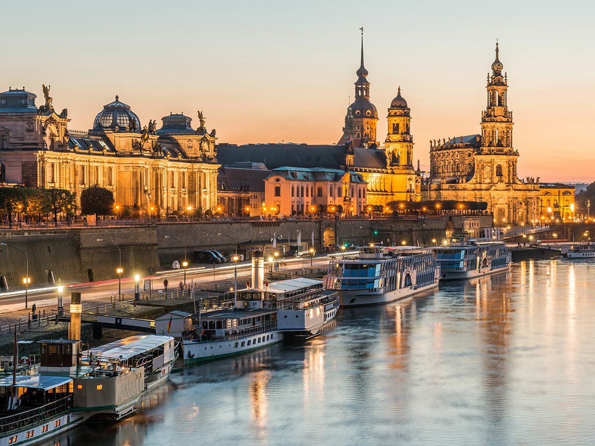 Dresden Altstadt bei Dämmerung mit beleuchteten Gebäuden wie Hofkirche und Residenzschloss am Elbufer, Schiffe im Fluss.