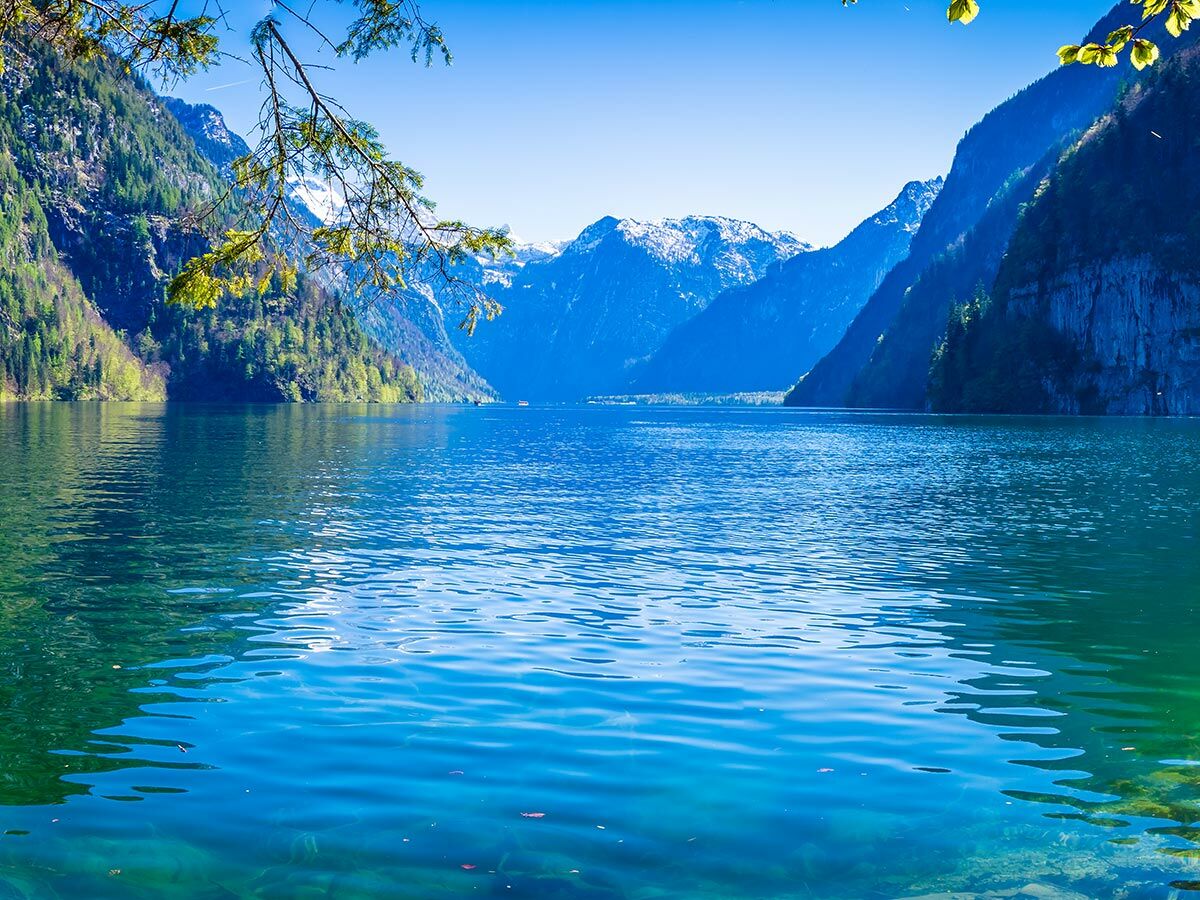 Malerischer Königssee mit klarem blaugrünem Wasser, umgeben von steilen, bewaldeten Alpenbergen und schneebedeckten Gipfeln unter blauem Himmel.