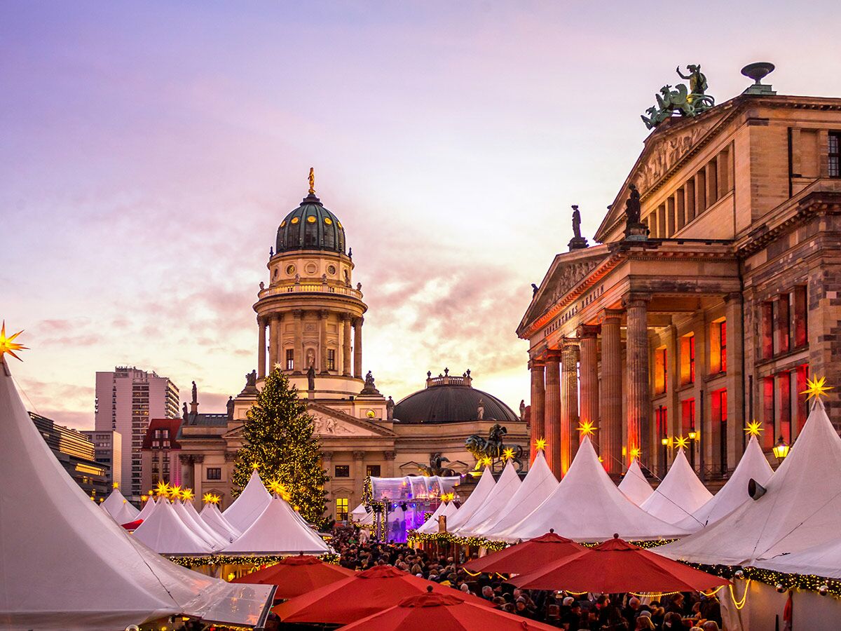 Weihnachtsmarkt Gendarmenmarkt Berlin bei Sonnenuntergang mit beleuchteten Ständen, festlichem Baum, Deutschem Dom und Konzerthaus.