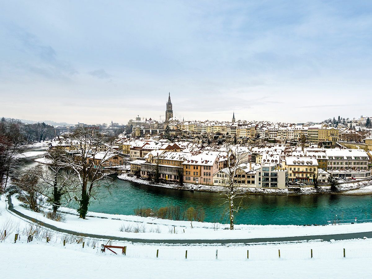 Panoramablick auf die schneebedeckte Stadt Bern im Winter, mit türkisfarbenem Aare-Fluss und dem Berner Münster.