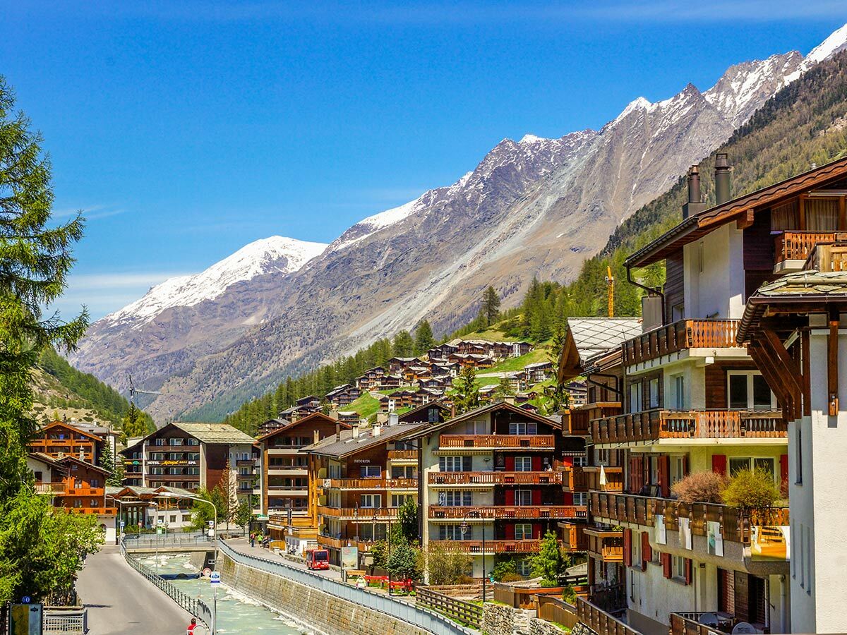 Panoramablick auf ein sonniges Alpendorf mit traditionellen Chalets, Fluss und majestätischen Schneebergen im Hintergrund.